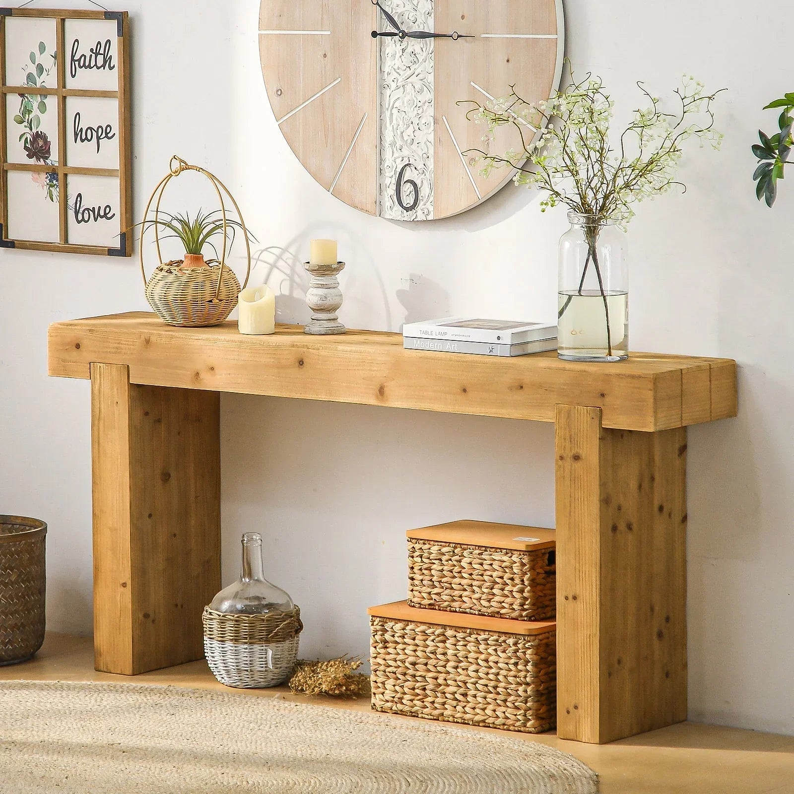 Solid wood console table styled with woven baskets, candle holders, greenery in a glass vase, wall clock, and farmhouse wall decor in a warm neutral entryway.
