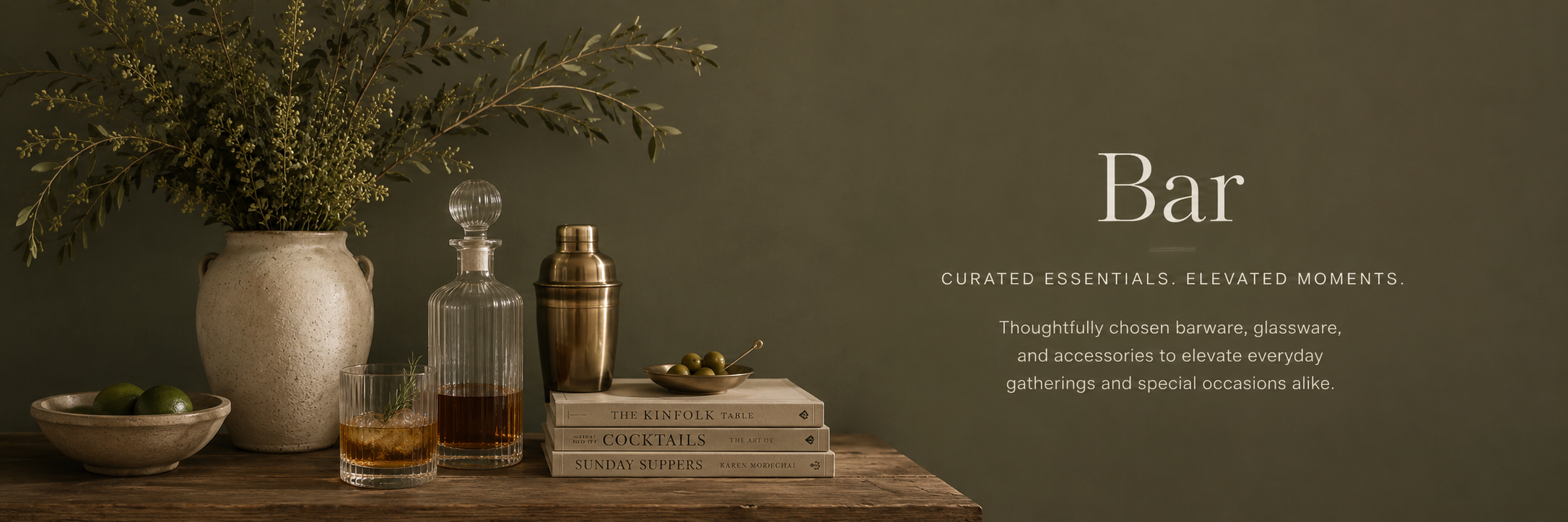 Warm editorial-style bar collection banner featuring refined glassware, brass cocktail accessories, soft botanicals, and moody olive tones styled in an elevated, intimate entertaining setting.