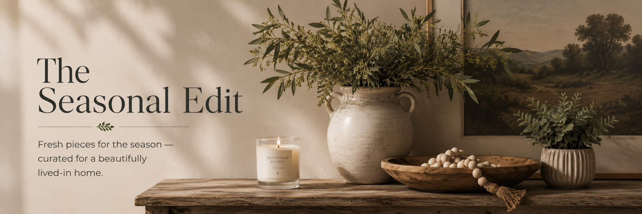 Neutral seasonal home decor banner featuring a rustic wood console styled with a ceramic vase of greenery, candle, wooden bowl, beads, potted plant, and landscape artwork beside text that reads The Seasonal Edit.